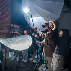 Students hold a light and reflector. A photographer holds their camera up to make the picture.