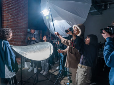 Students hold a light and reflector. A photographer holds their camera up to make the picture.