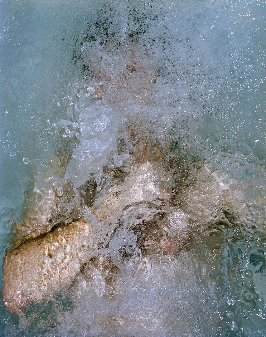 Light blue, bubbling water obscures the body (torso up) of a white person lying on their back with their arm bent over their chest. Their face is obscured by the rushing water. They appear to be falling backward.