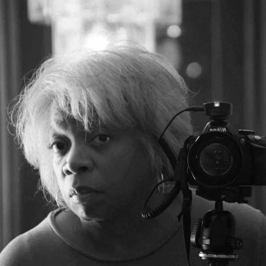 Image Description: A black and white selfie of Karen D. Bailey. Karen's face is next to her camera which is set up on a tripod to make the image.