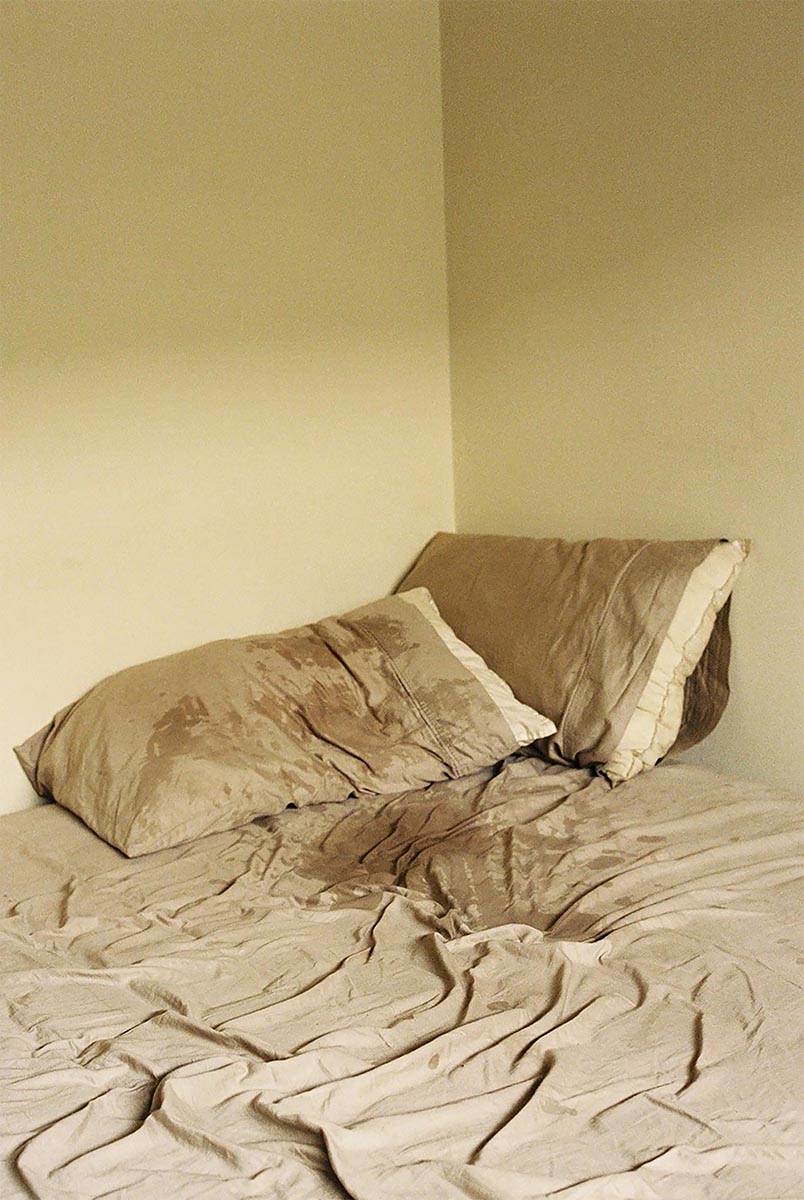Two pillows partially covered with tan pillow cases sit atop wrinkled, sweat-soaked tan sheets. A light yellow wall completes the background. The bed appears to have been slept in, The soft light projected in the image gives the room a warm glow.