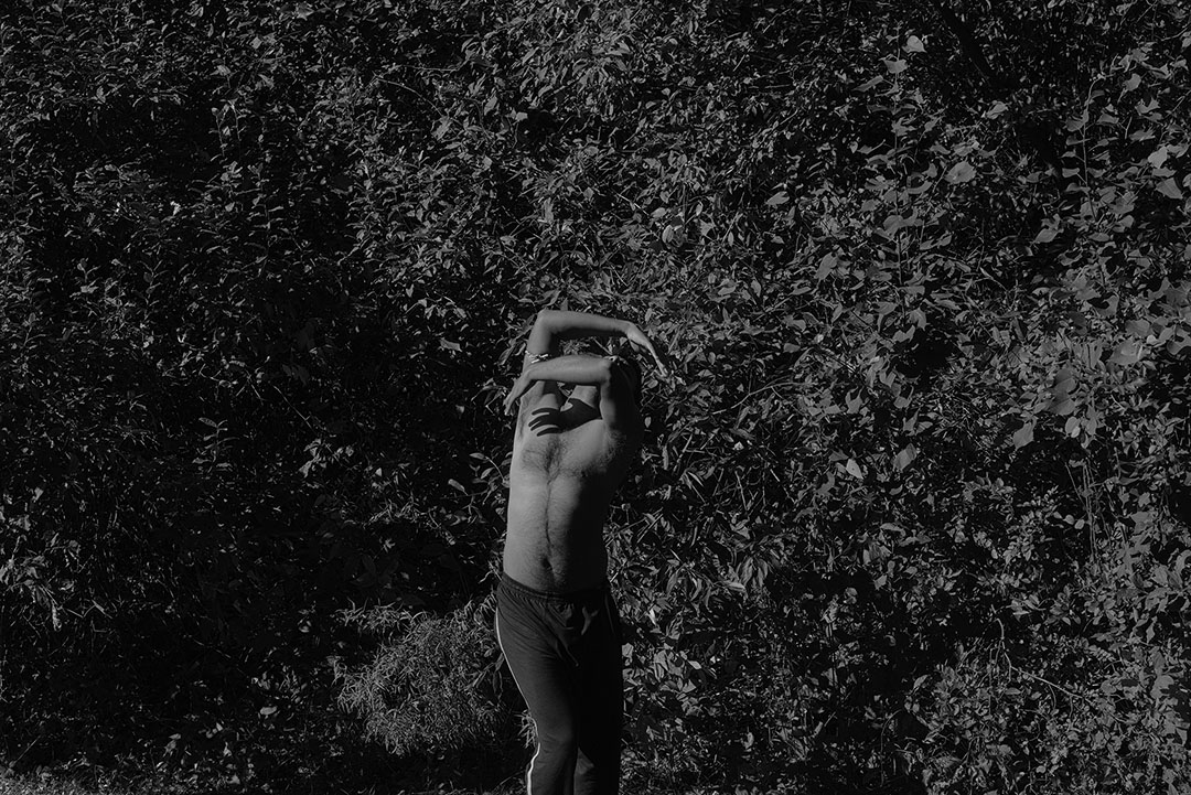 Black and white image of a shirtless masculine person who presents as South Asian, wearing dark colored track pants with a white strip along the side. Their body is slightly contorted, arms cradled above their head as it is tilted backwards positioned towards the sun light. Behind the subject is a lush, dense background of various foliage.