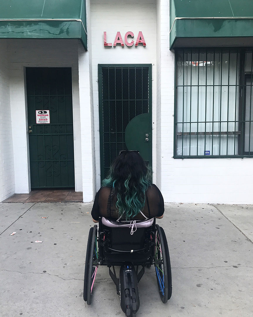 A woman sits in a wheelchair on a sidewalk with her back to the camera. Her long wavy hair capped at its ends by blue highlights, contrasts with the pink fabric slightly showing underneath her mesh top. A pair of sunglasses rests on the top of her head. The front of her body is facing the door of a white building adorned with green awnings. A red sign with the letters “LACA” capitalized sits just above the entryway. Flanking the entrance is another door to the left and a window to the right. All entry points are covered with bars.
