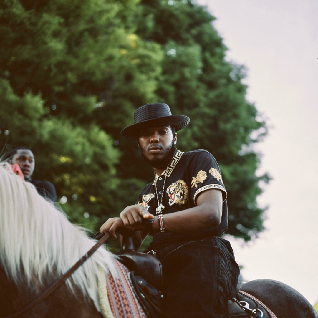 Black male riding a dark colored horse with a white mane. The subject is wearing a black cowboy hat, black pants, and a short sleeve shirt with a tiger on the front and yellow roses on the sleeve. Behind the man is a large tree with green leaves and another Black male in the background.