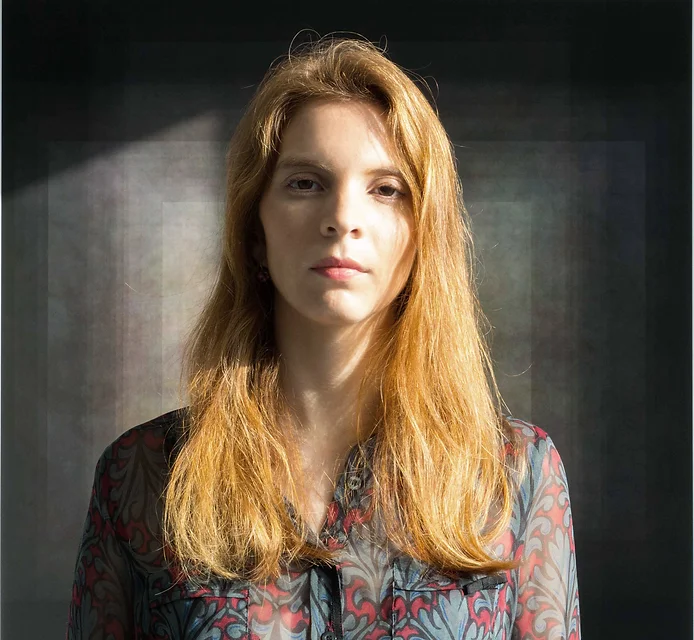 Portrait of a woman of European descent with long reddish-orange hair. She is wearing a red blouse with large bluish-gray leaves scattered about. The colors on the garment are dull. In the background is a series of rectangles in various shades of gray. The objects start out small, becoming bigger and bigger as they shift from light to dark. 