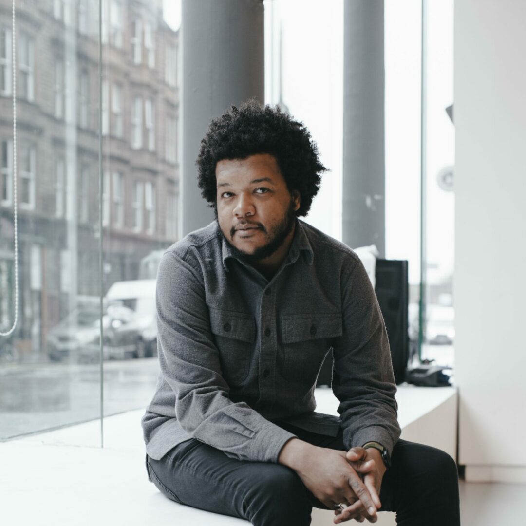 A portrait of Chicago based filmmaker and curator Amir George. They are wearing a grey button up with black pants and sit on a long ledge by the window as they look into the camera.